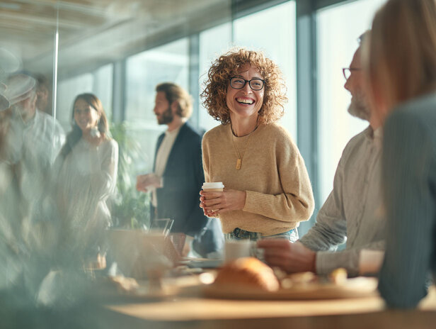 Usměvavá žena v příjemném svetru drží kávu a užívá si živé obchodní schůzky s kolegy ve světlé kanceláři.