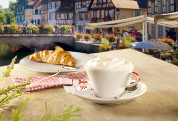 Lahodná mléčná káva a croissant se podávají s malebnými historickými domy v pozadí, ideální pro odpočinkové ráno.