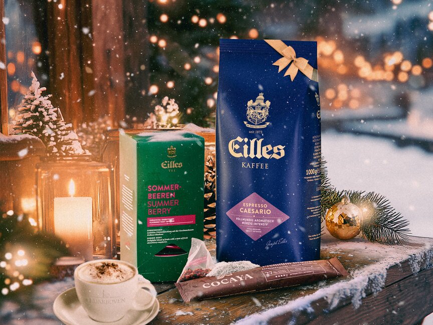 Šálek teplé kávy Eilles se svátečním čajem, čokoládou a zimní výzdobou na zasněženém stole, ideální pro příjemnou sváteční atmosféru.