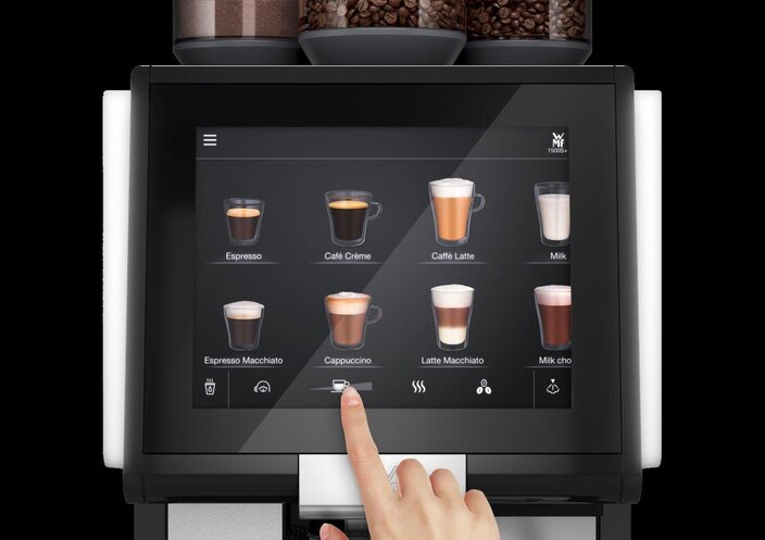 Interaktivní kávovar s dotykovou obrazovkou zobrazující různé nápoje, například cappuccino a latté.