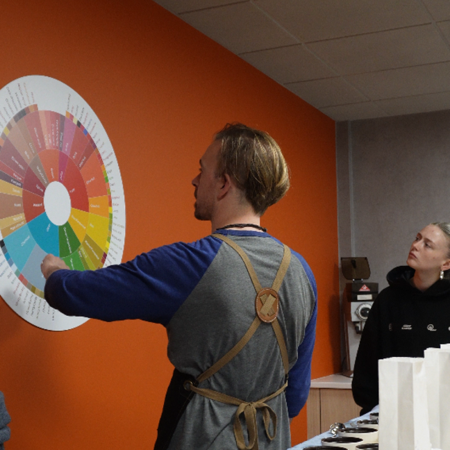 A coffee expert explains flavour profiles using a colour wheel, guiding keen learners in a vibrant café setting.