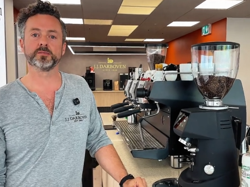 Barista at a coffee shop stands next to an espresso machine and grinder, highlighting artisanal coffee preparation.