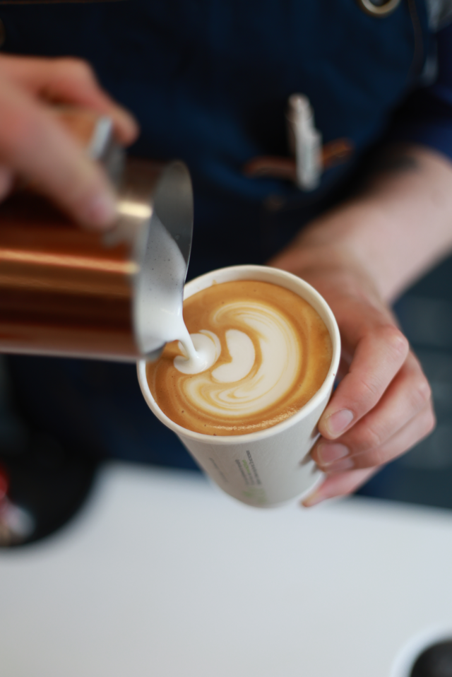Barista pouring latte art into a takeaway cup, showcasing creamy milk foam and rich coffee. Perfect morning brew.