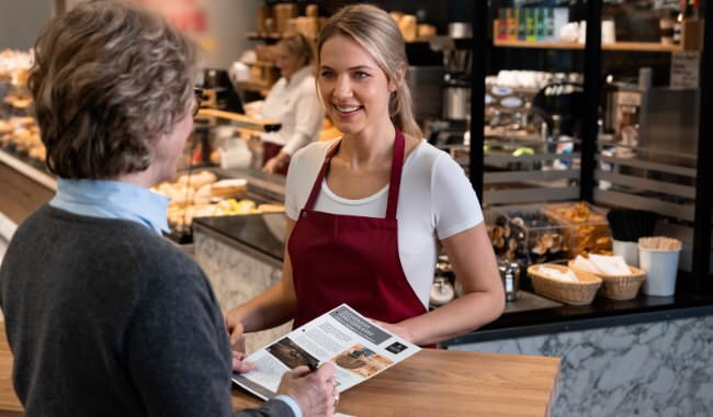 Customer being served at a cafe by a smiling barista in a maroon apron, showcasing a friendly atmosphere.