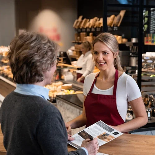 Friendly bakery staff member assisting a customer with a menu in a warm, inviting café environment.