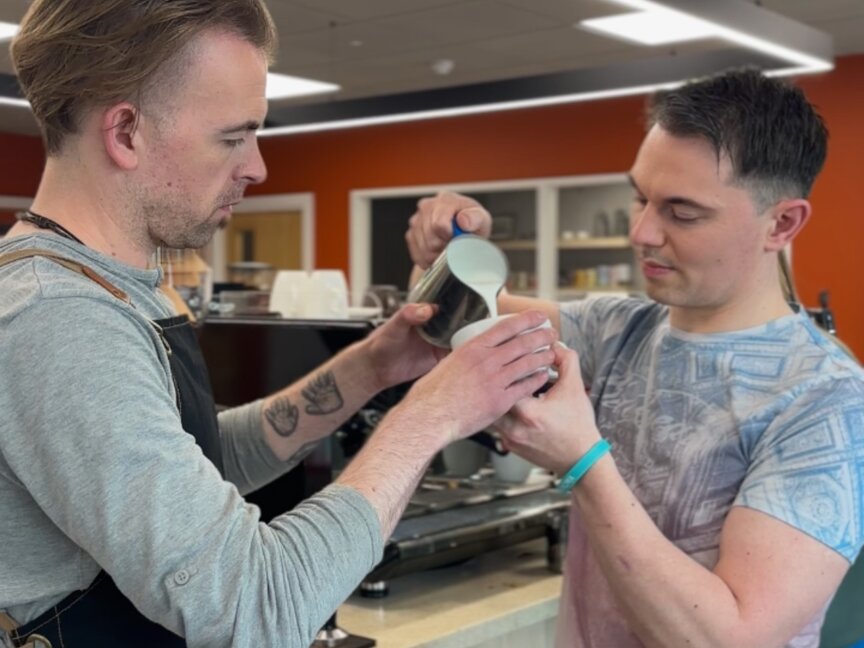 Two baristas collaborating to pour milk into a cup, showcasing coffee art techniques in a modern café setting.