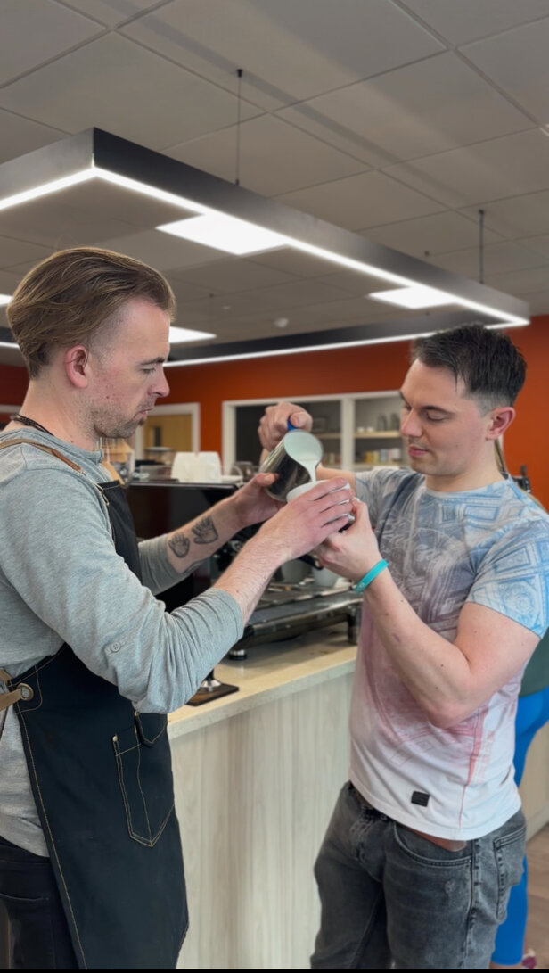 Two baristas collaborating to pour milk into a cup, showcasing coffee art techniques in a modern café setting.