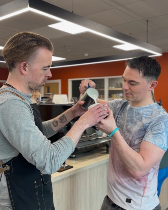 Two baristas collaborating to pour milk into a cup, showcasing coffee art techniques in a modern café setting.