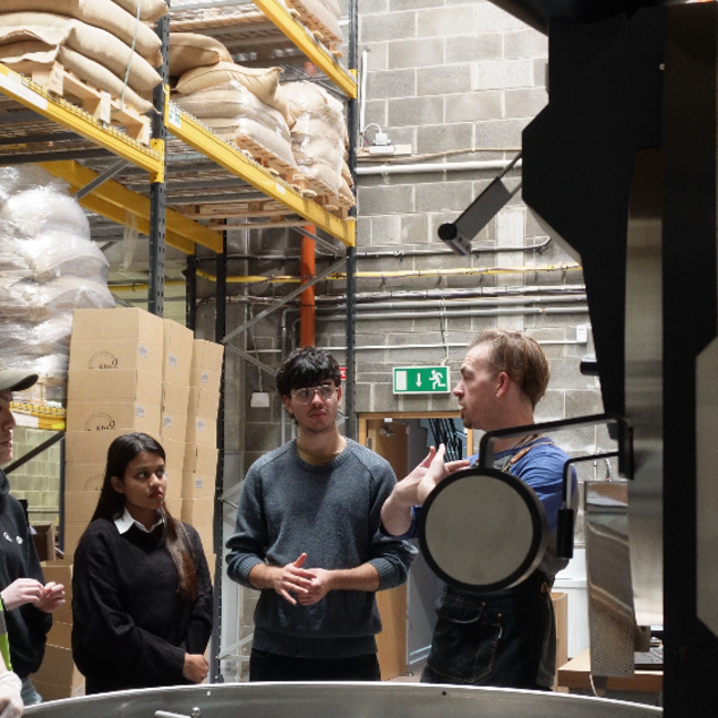 Coffee roasting workshop with participants learning from an expert in a warehouse environment, surrounded by equipment and supplies.
