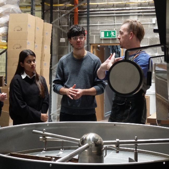 Coffee roasting workshop with participants learning from an expert in a warehouse environment, surrounded by equipment and supplies.