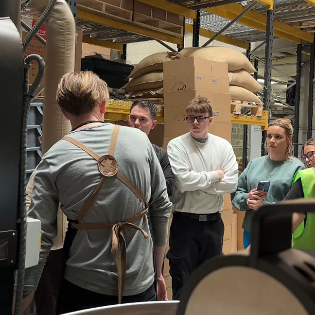 Coffee roasting demonstration with attendees observing in a warehouse, surrounded by sacks of raw coffee beans.