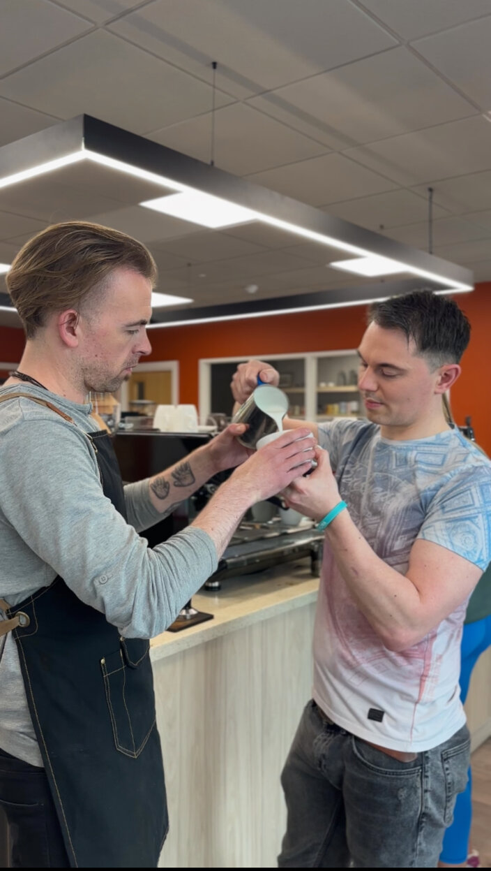Two baristas demonstrate latte art techniques in a vibrant café setting, showcasing skills with milk and coffee.