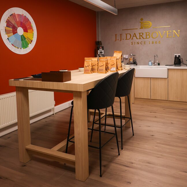 Stylish café interior featuring a modern wooden table, seating, and J.J. Darboven coffee bags against a vibrant orange wall.