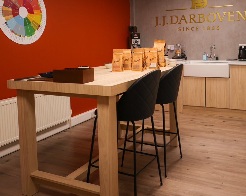 Stylish café interior featuring a modern wooden table, seating, and J.J. Darboven coffee bags against a vibrant orange wall.