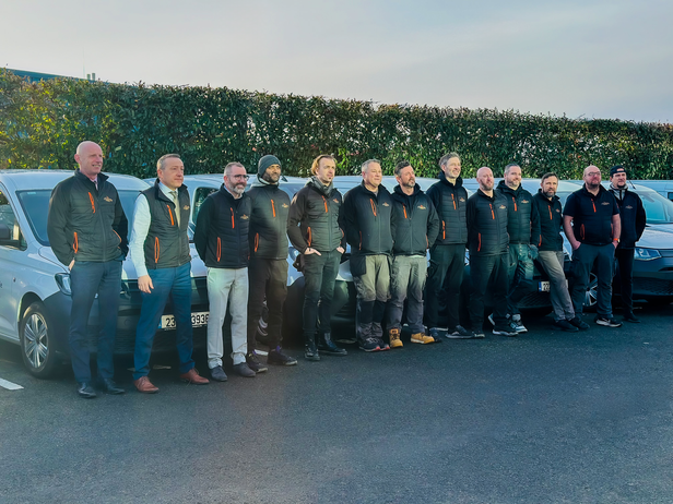 Team of professionals in branded jackets posing with company vehicles, showcasing unity and teamwork in a commercial setting.