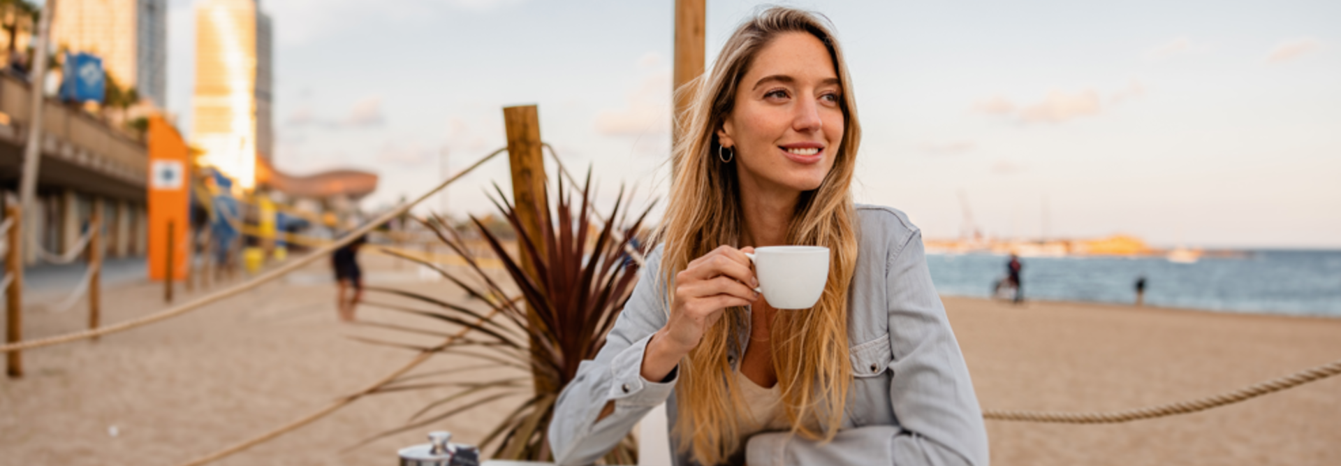Femme détendue dégustant un café sur une plage de sable avec l'horizon de la ville en arrière-plan. Parfait pour les cafés côtiers.