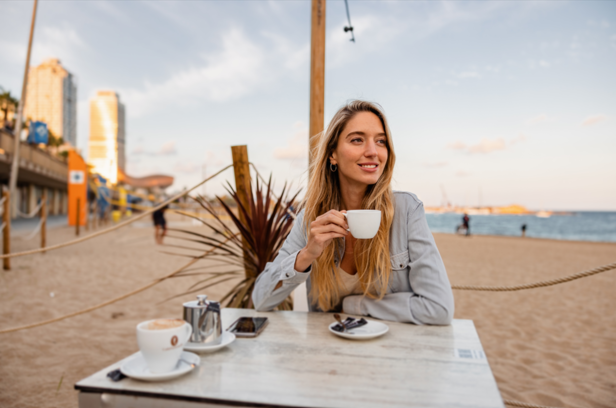Entspannende Frau genießt Kaffee an einem Sandstrand mit der Skyline der Stadt im Hintergrund. Perfekt für Küsten-Café-Vibes.