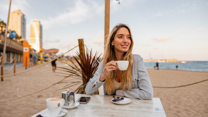 Relaxing woman enjoying coffee on a sandy beach with city skyline in the background. Perfect for coastal café vibes.