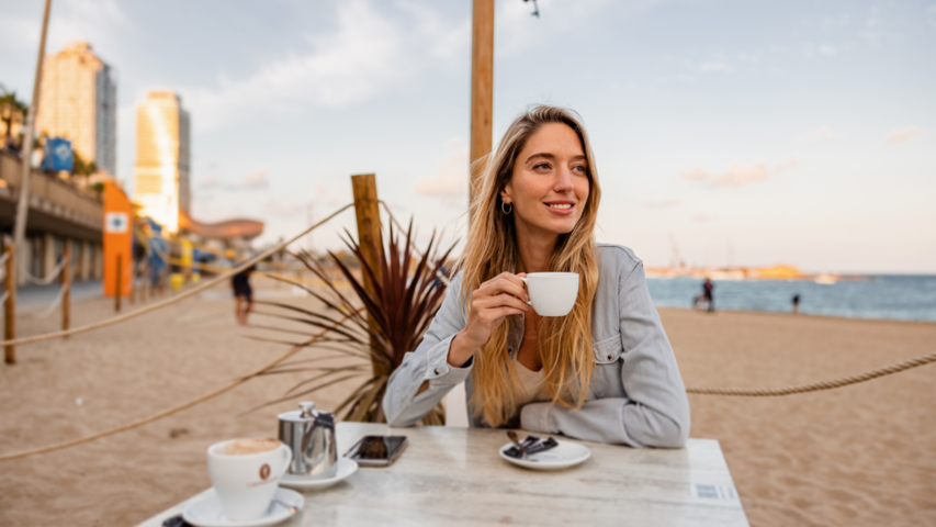Relaxing woman enjoying coffee on a sandy beach with city skyline in the background. Perfect for coastal café vibes.