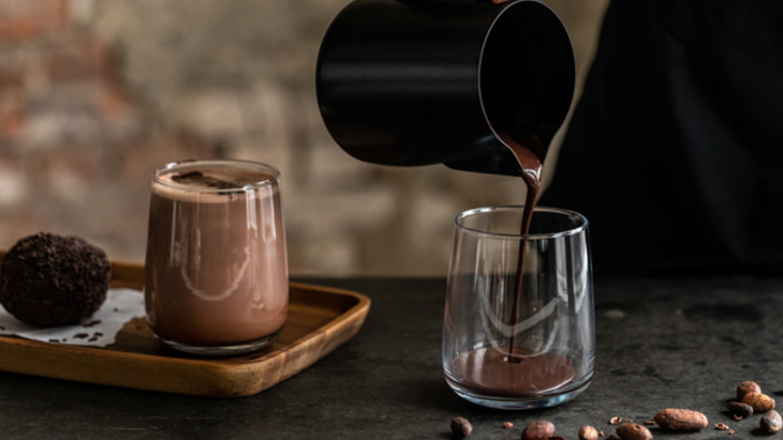 Un riche chocolat chaud est versé dans un verre à partir d'une cruche noire, accompagné de fèves de cacao et d'une truffe au chocolat sur un plateau en bois.