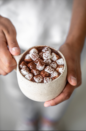 Cacao chaud garni de mini-guimauves et de poudre de cacao, tenu par une personne dans une tasse texturée. Une gâterie hivernale parfaite.