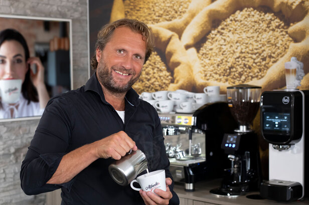 Barista schenkt vakkundig melk in een koffiekopje in een modern café, met koffieapparatuur en gebrande bonen op de achtergrond.