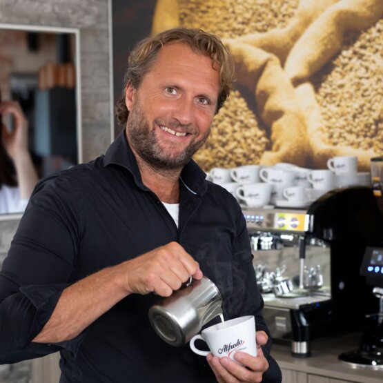 Barista schenkt vakkundig melk in een koffiekopje in een modern café, met koffieapparatuur en gebrande bonen op de achtergrond.
