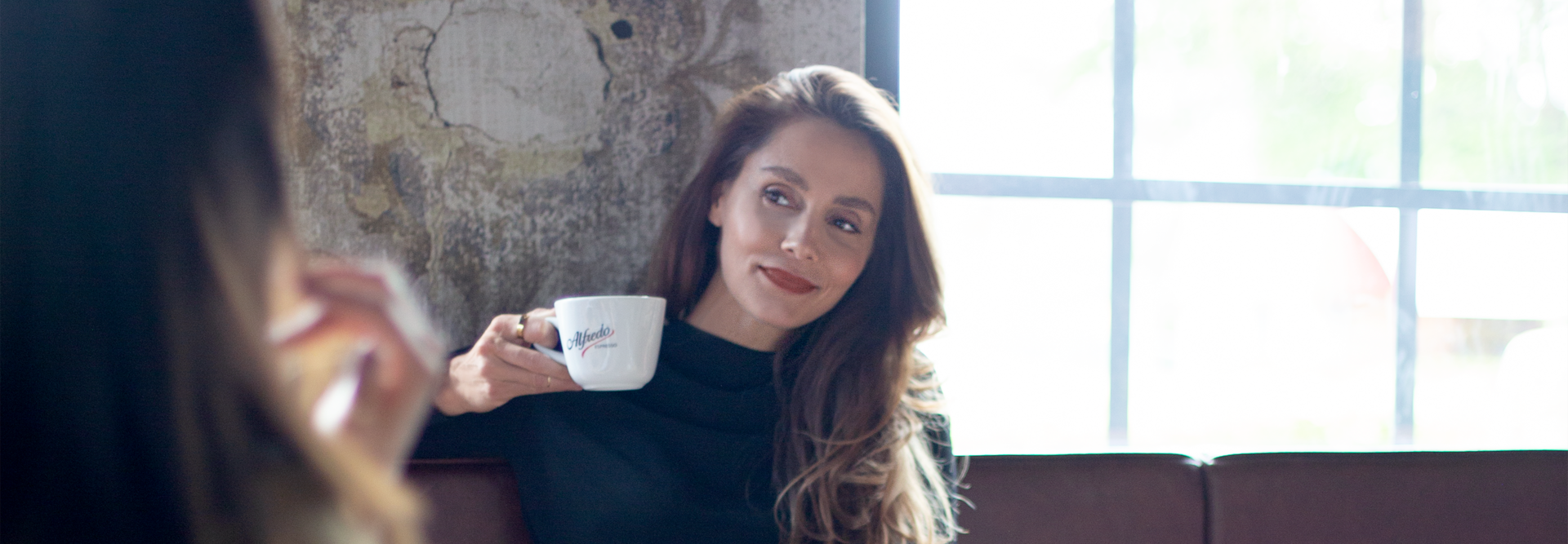 Vrouw geniet van koffie in een café, draagt een stijlvolle zwarte jurk, met een ontspannen glimlach en een modern interieur op de achtergrond.