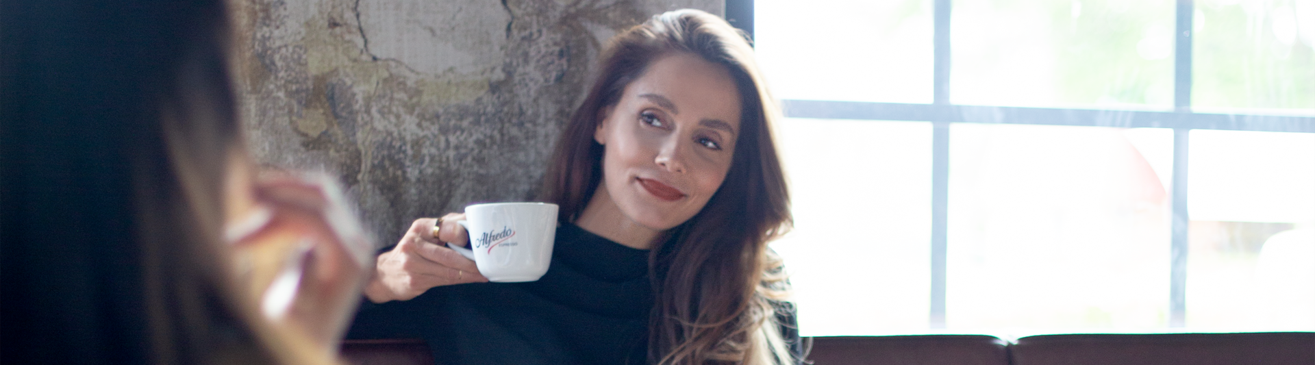 Vrouw geniet van koffie in een café, draagt een stijlvolle zwarte jurk, met een ontspannen glimlach en een modern interieur op de achtergrond.
