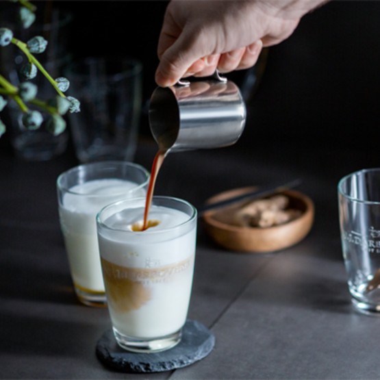 Barista schenkt koffie met opgeschuimde melk in elegante glazen, naast koekjes, op een stijlvol donker aanrecht.