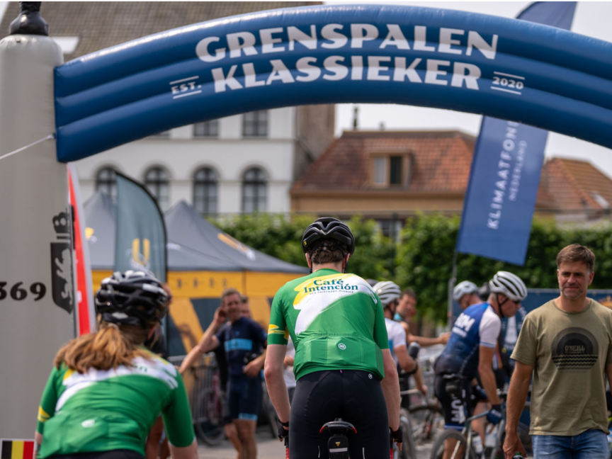 Fietsers op het evenement Grenspalen Klassieker, met Café Intención, met een feestelijke sfeer en levendige spandoeken.