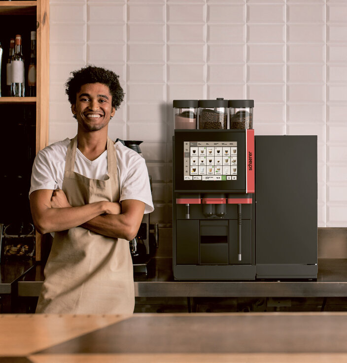 Een glimlachende barista in een schort staat zelfverzekerd naast een moderne koffiemachine in een stijlvol café.