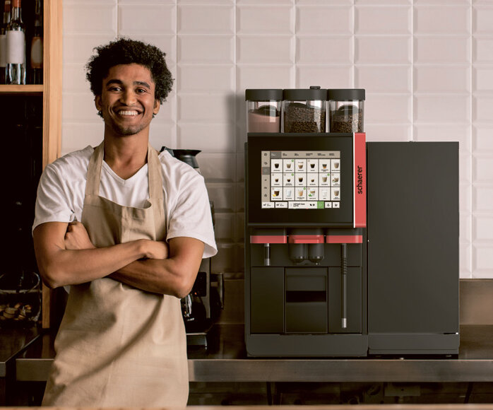 Een glimlachende barista in een schort staat zelfverzekerd naast een moderne koffiemachine in een stijlvol café.