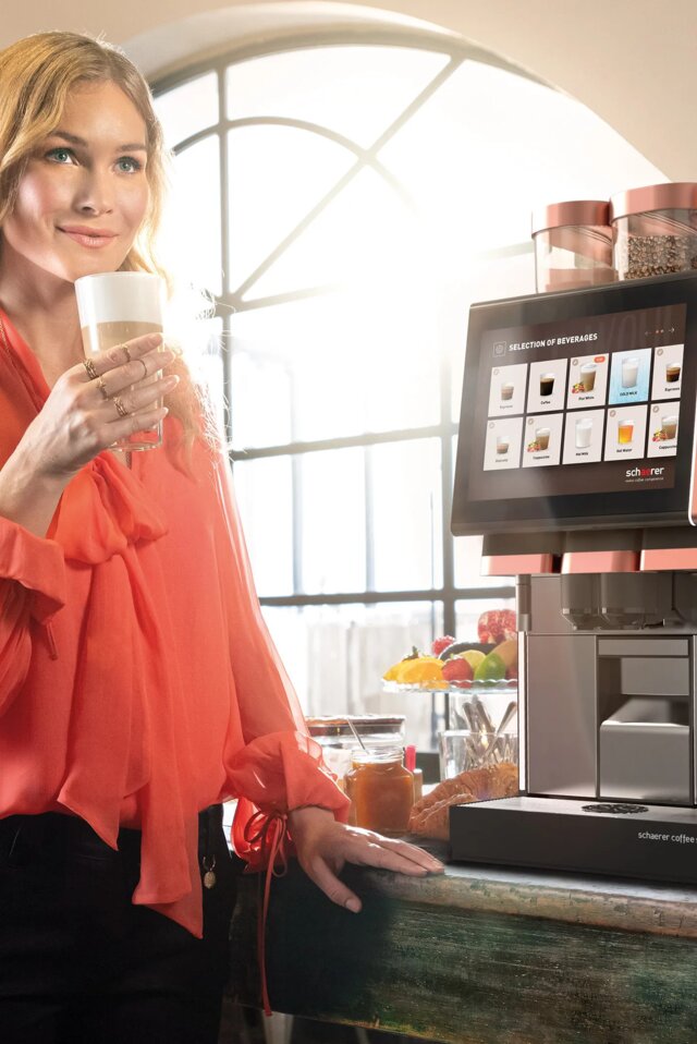 Een vrouw in een kleurrijke blouse geniet van een koffiedrankje naast een strak ogende drankautomaat in een stijlvol café.