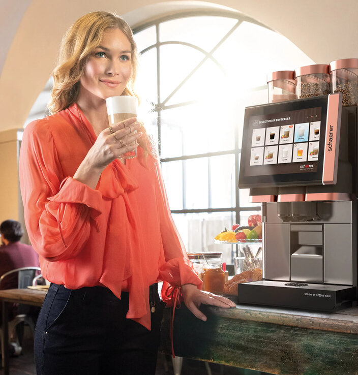 Een vrouw in een kleurrijke blouse geniet van een koffiedrankje naast een strak ogende drankautomaat in een stijlvol café.