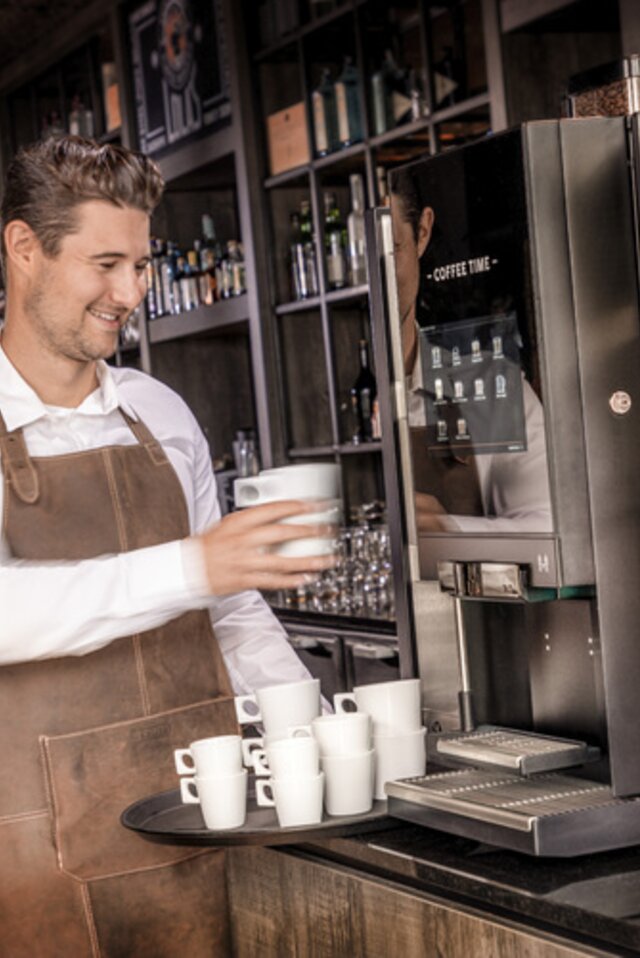 Een barista die koffie zet met een moderne machine, omringd door kopjes, in een stijlvolle caféomgeving.