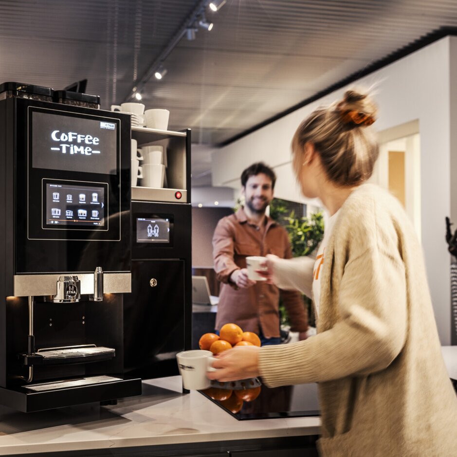 Een moderne kantoorkeuken met een koffiezetapparaat en twee collega’s die van een pauze genieten en samen koffie en verse sinaasappels eten.