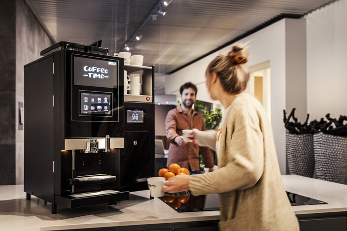 Een moderne kantoorkeuken met een koffiezetapparaat en twee collega’s die van een pauze genieten en samen koffie en verse sinaasappels eten.