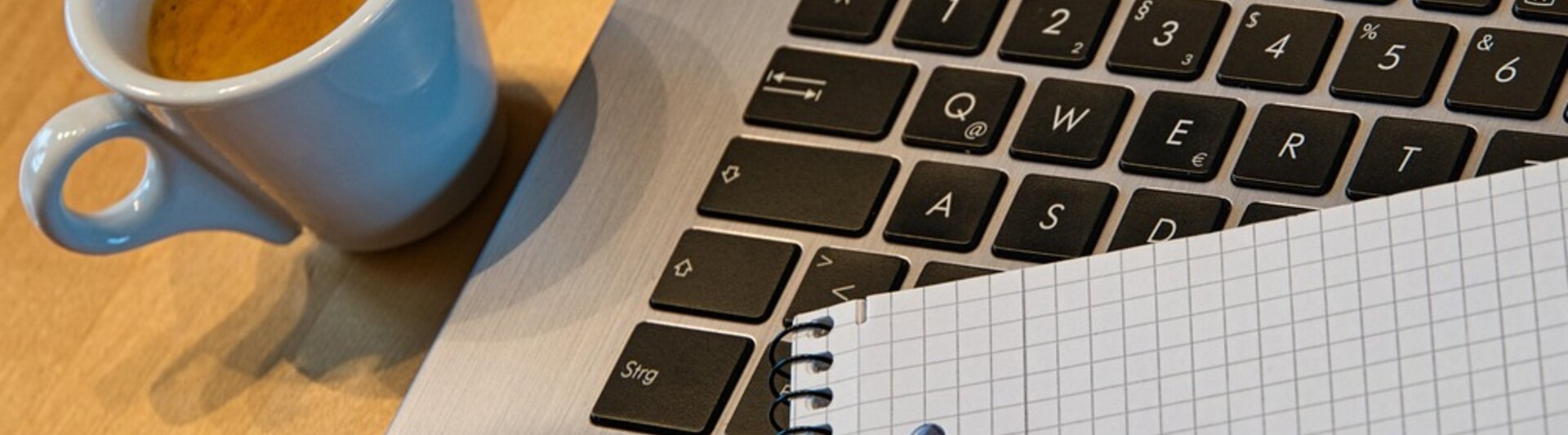 Laptop obok filiżanki kawy, notatnika i długopisu na drewnianym biurku – idealne rozwiązanie do pracy w domowym biurze.