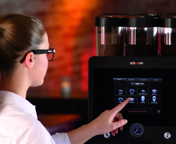 Kobieta korzystająca z ekranu dotykowego ekspresu do kawy Schaerer do wyboru opcji kawy w stylowej kawiarni.