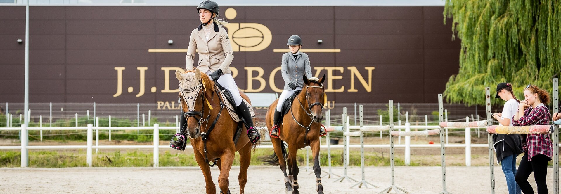 Dwóch jeźdźców na koniach uczestniczy w treningu w ośrodku jeździeckim J.J. De Boeren, obserwowanym przez widzów.