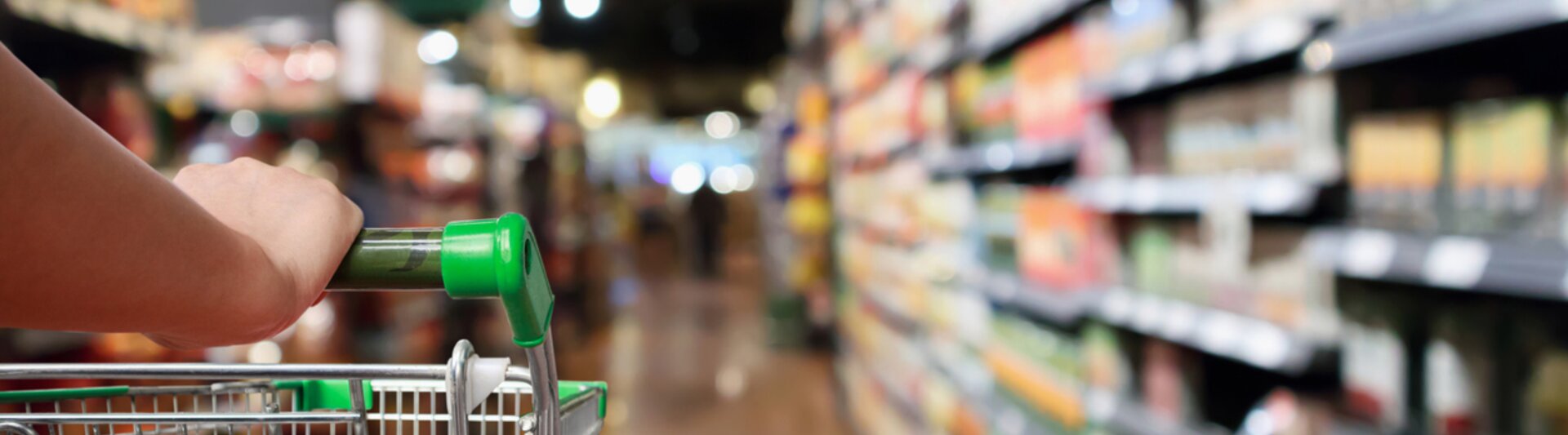 Wózek sklepowy w alejce sklepu spożywczego, otoczony półkami z różnymi produktami i delikatnym oświetleniem.