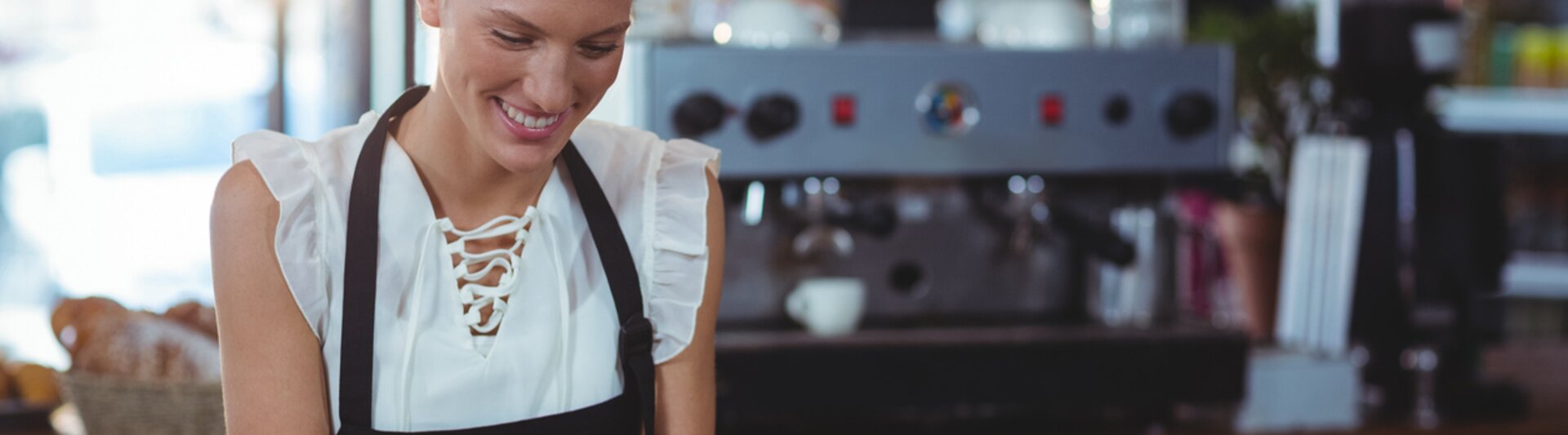 Baristka korzystająca z tabletu w kawiarni, uśmiechająca się, gdy obsługuje zamówienia za drewnianą ladą, a w tle widać ekspres do kawy.