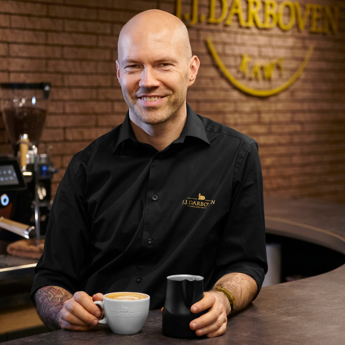 Uśmiechnięty barista obok latte i dzbanka z mlekiem, prezentujący doświadczenie w akademii kawy J.J. Darboven w stylowym otoczeniu.