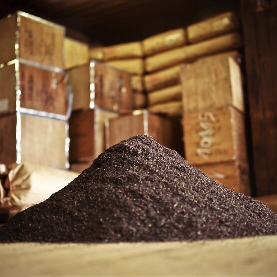 Mountain of dark ground spices in a rustic storage area with stacked boxes in the background.