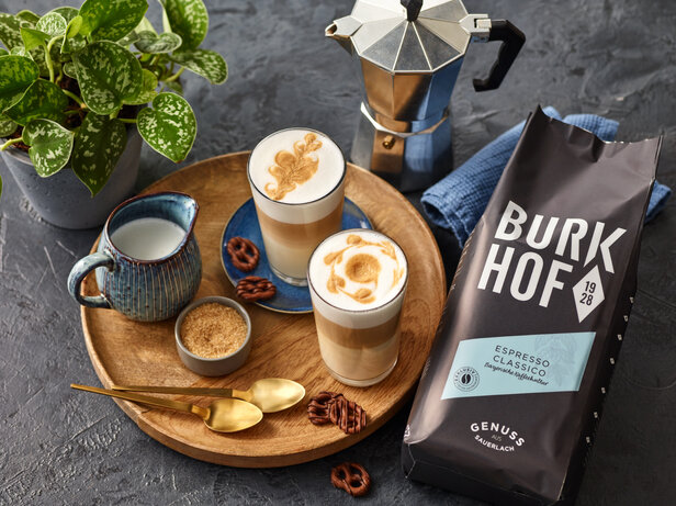 Latte Macchiato, serviert mit Burkhof Kaffee, Zucker und Brezeln, ergänzt durch eine üppige Grünpflanze im Hintergrund.