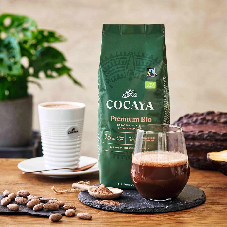 Kakao Cocaya Premium Bio, čerstvý nápoj, kakaové boby a rostlina na dřevěném stole – to vše podtrhuje myšlenku udržitelného požitku.