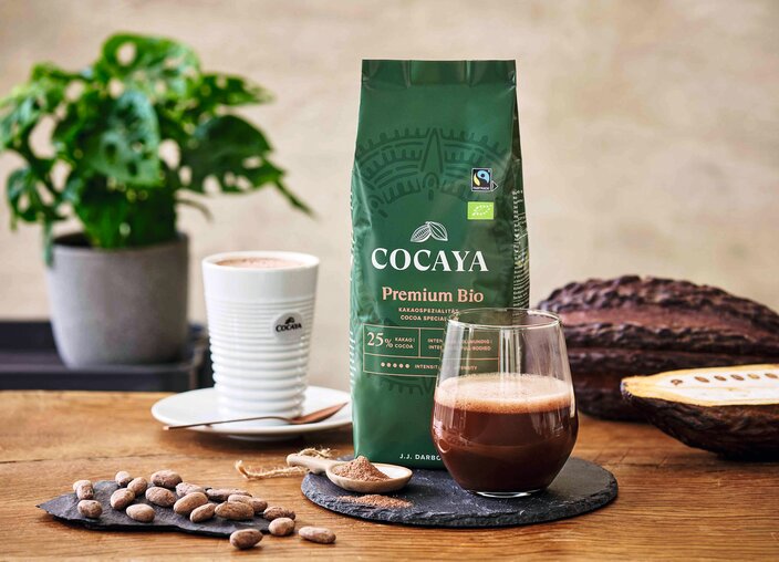 Kakao Cocaya Premium Bio, čerstvý nápoj, kakaové boby a rostlina na dřevěném stole – to vše podtrhuje myšlenku udržitelného požitku.
