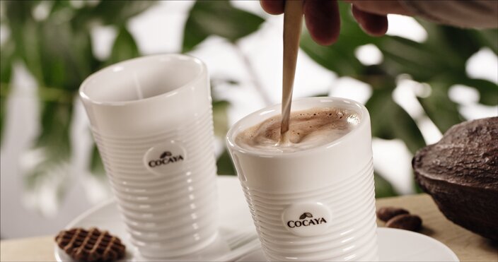 Steaming cocoa being stirred in a Cocaya mug, paired with chocolate wafers, set against lush greenery.