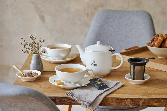 Elegant Eilles tea set on a wooden table, featuring tea cups, a teapot, and snacks for a delightful afternoon tea.